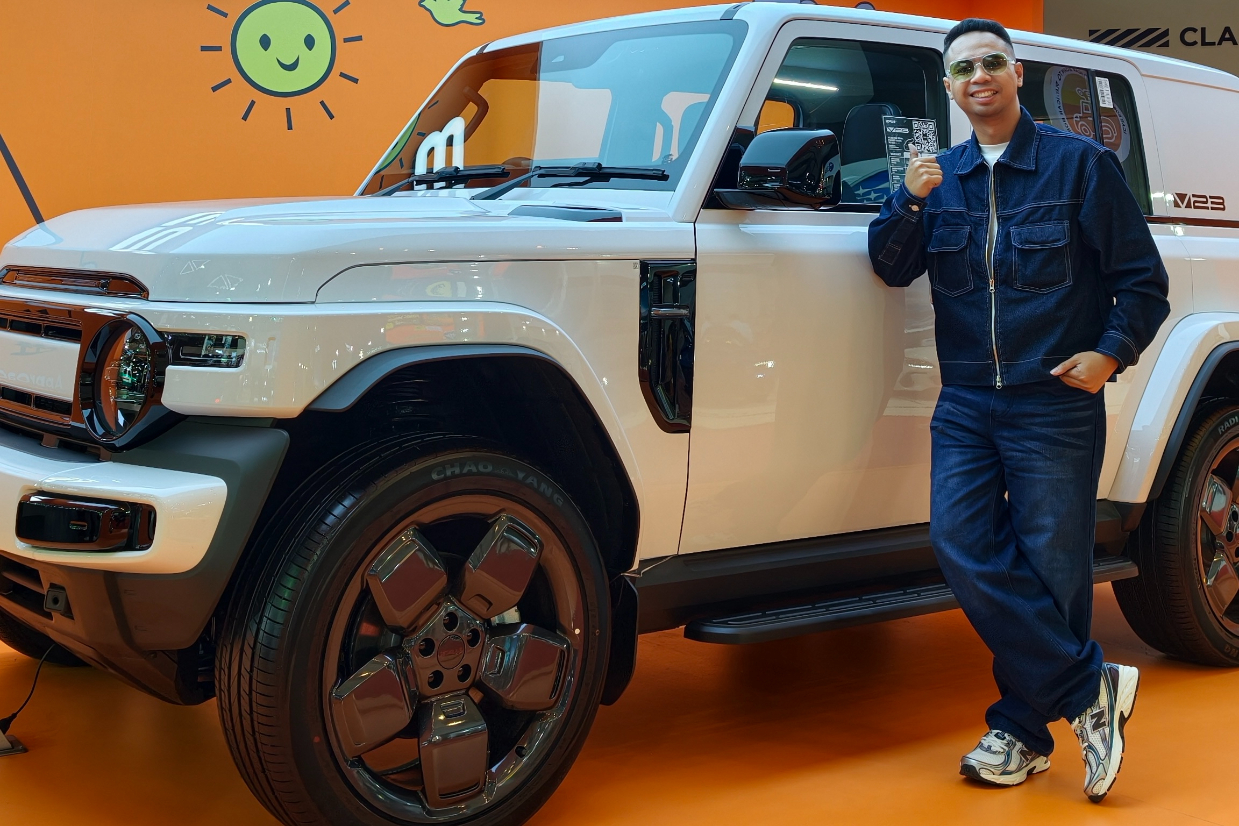A person standing beside a white SUV against a vibrant orange background with graphics. The individual is wearing a denim outfit and stylish sunglasses, holding a card.