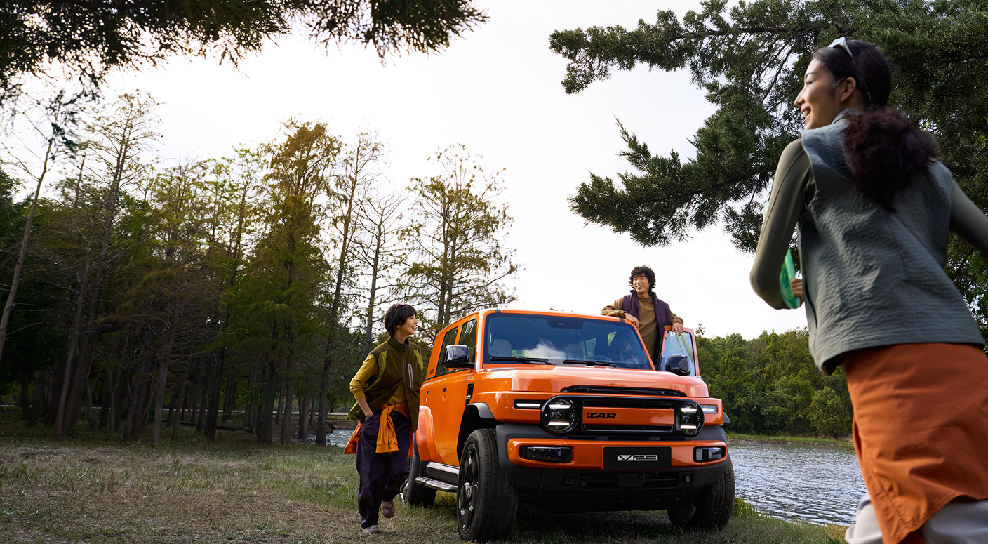 A group of three people enjoying a day outdoors near a lake, gathered around an orange off-road vehicle. Trees and greenery are visible in the background.